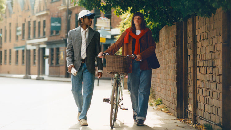 Two people walking and talking on a city sidewalk beside a brick wall, with one person pushing a bicycle. The person on the left wears a gray blazer, jeans, and a white cap, while the person on the right wears a brown coat, a bright red scarf, and a dark beanie.