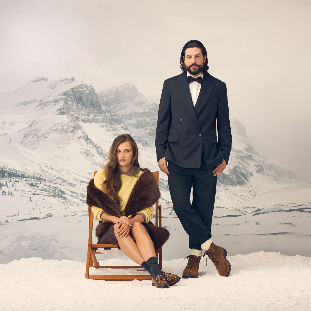A man standing next to a woman sitting down against a snowy backdrop, the man is wearing a pair of brown boots, and the woman is wearing a pair of brown loafers