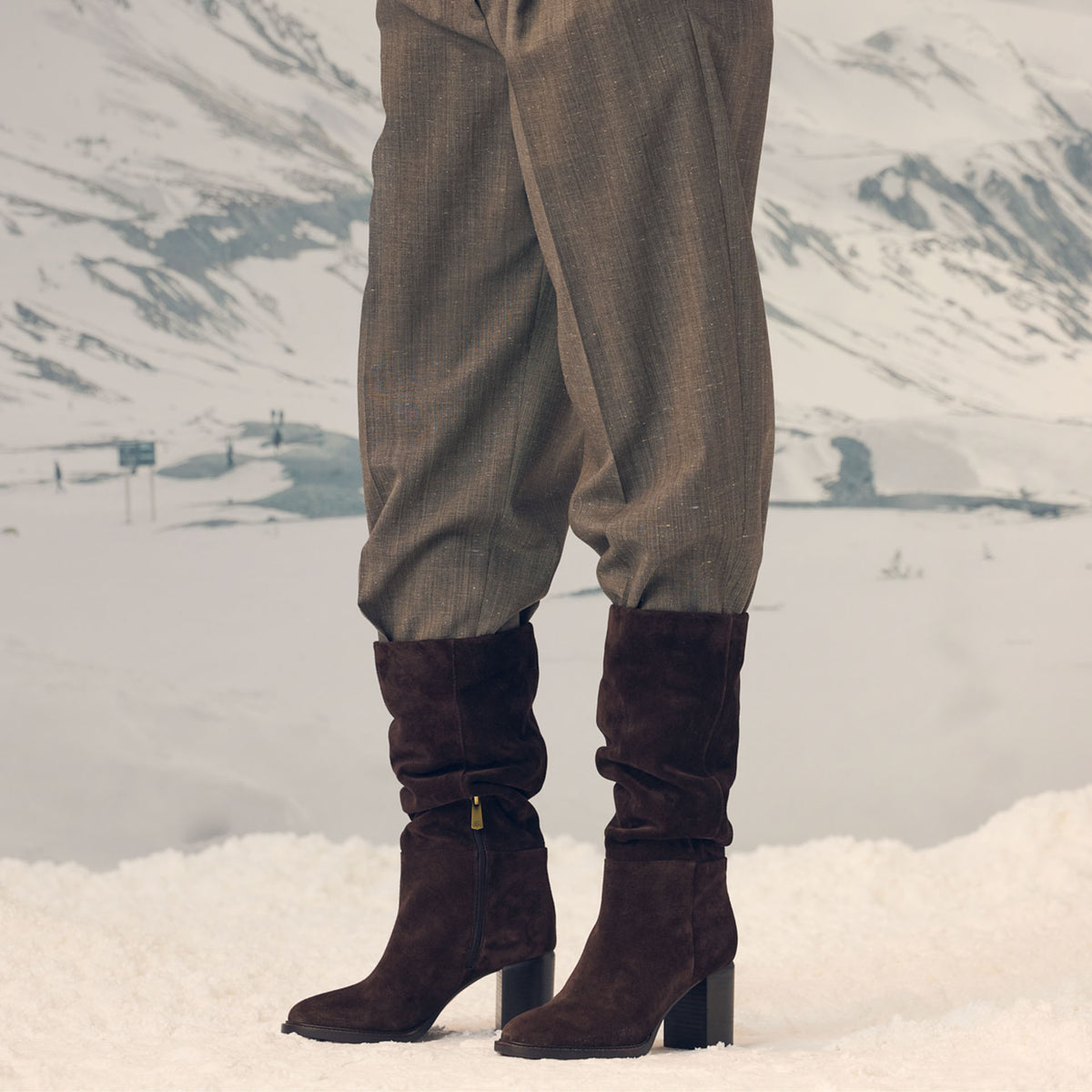 A close-up shot of a woman wearing a pair of tall brown suede heeled boots with pants tucked into them against a snowy backdrop