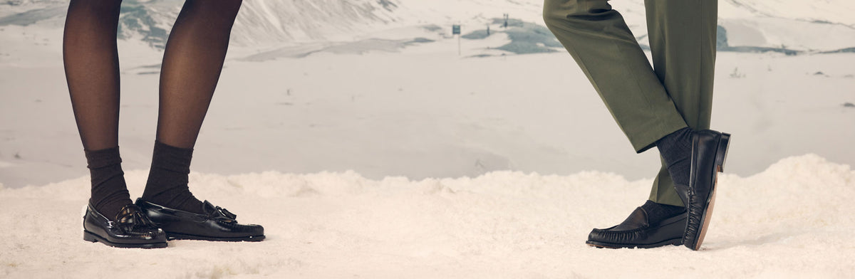 A cropped outdoor winter scene showing the lower legs of two people standing in snow, both wearing black loafers.