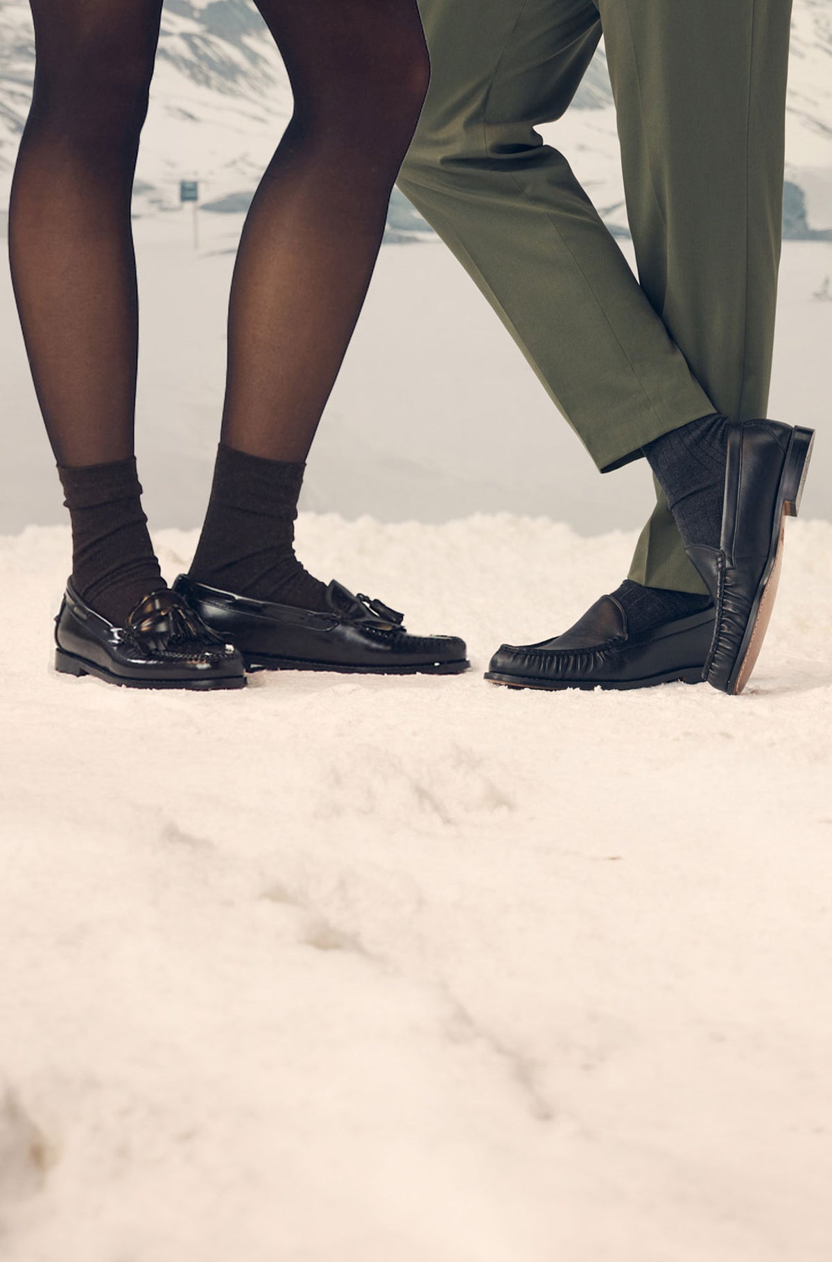 A cropped outdoor winter scene showing the lower legs of two people standing in snow, both wearing black loafers.