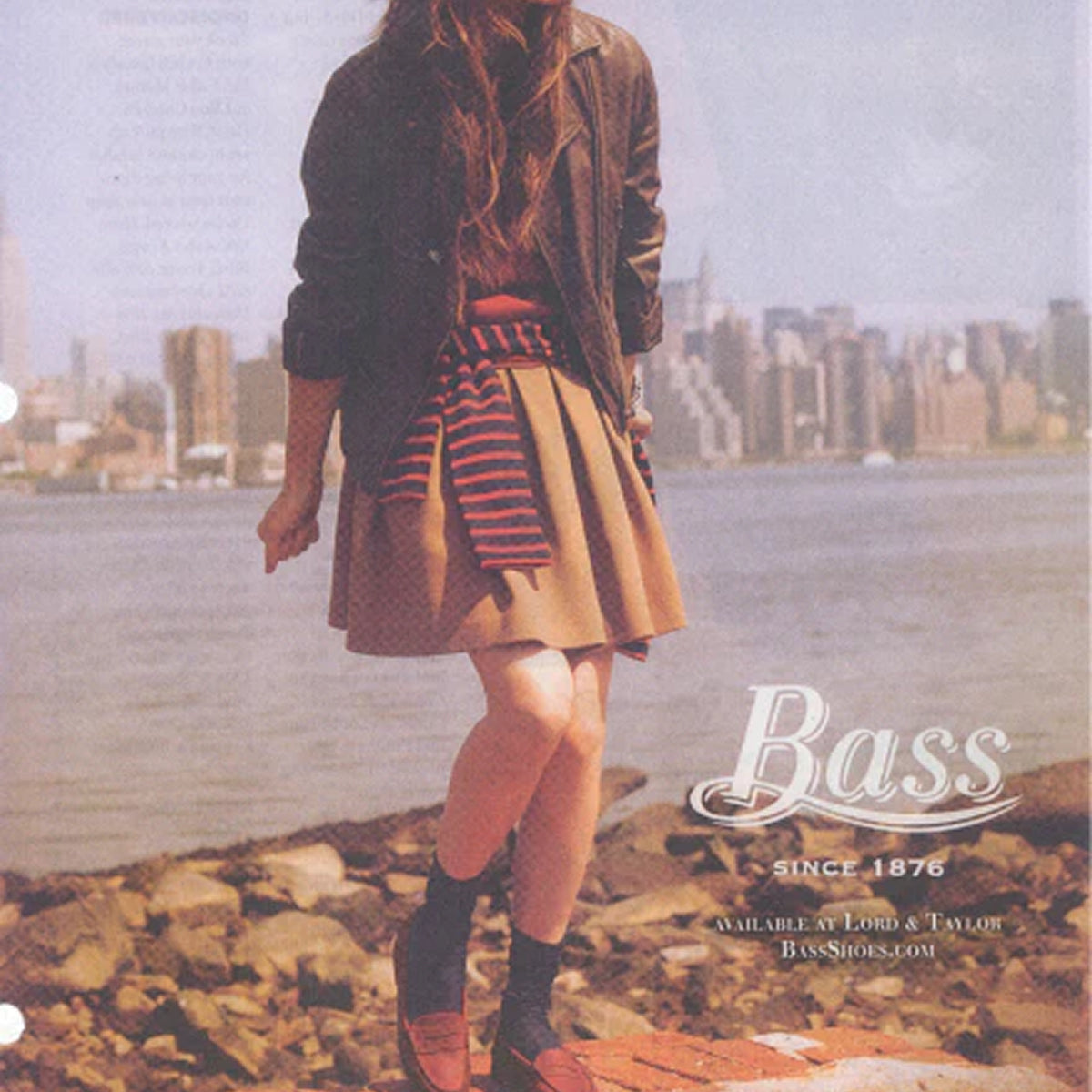 A girl standing on rocks by the water with the city skyline behind her wearing a beige skirt and brown loafers