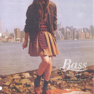 A girl standing on rocks by the water with the city skyline behind her wearing a beige skirt and brown loafers