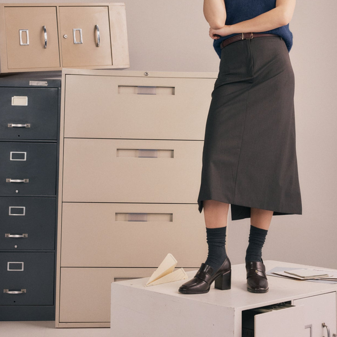 A woman standing on a file cabinet wearing a dark grey skirt with wine-colored heeled loafers