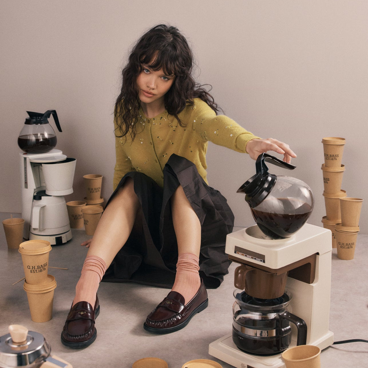 Woman sitting on the floor wearing wine-coloured loafers tilting a coffee machine and surrounded by coffee cups