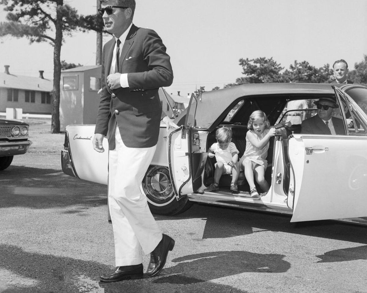 A black and white image of a man stepping out of a white car with children sitting in the back seat